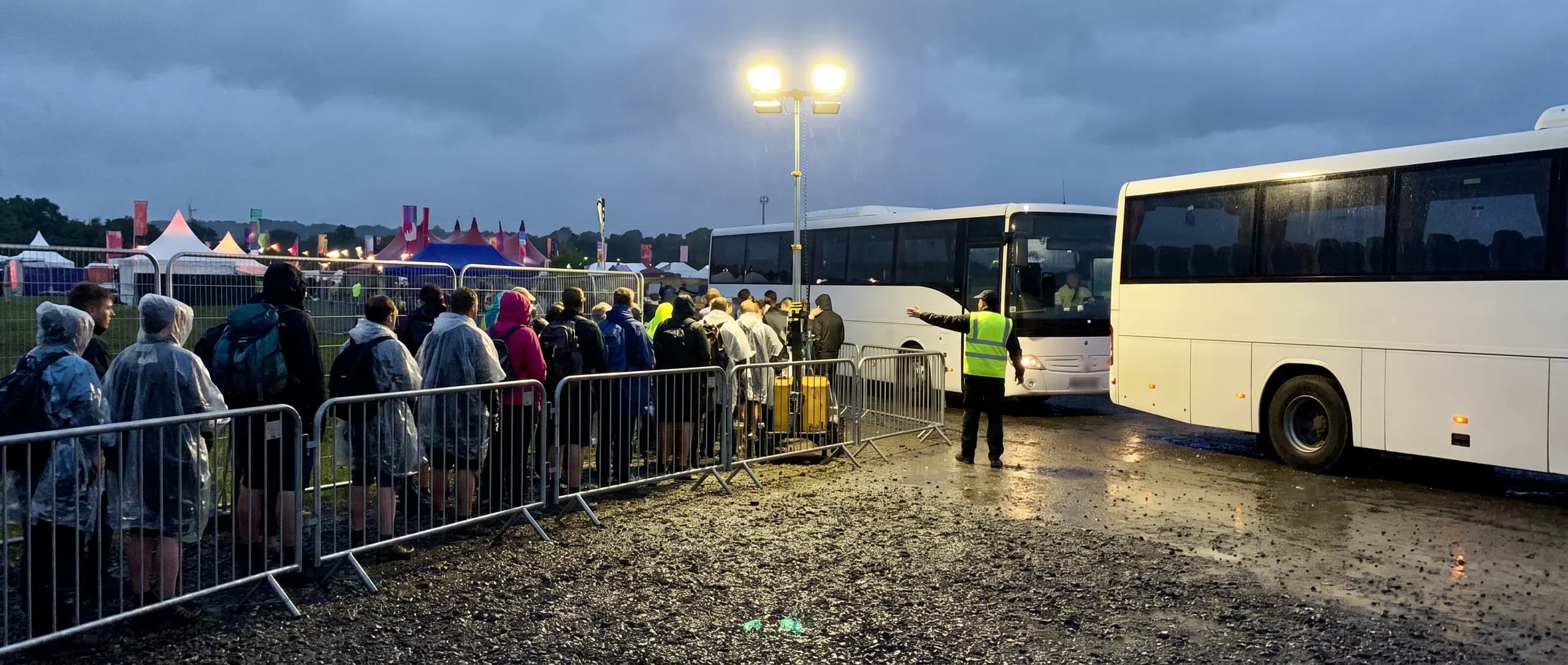 Festivalbezoekers in regenponcho's staan na afloop achter dranghekken in de rij voor twee pendelbussen, een verkeersregelaar wijst de weg