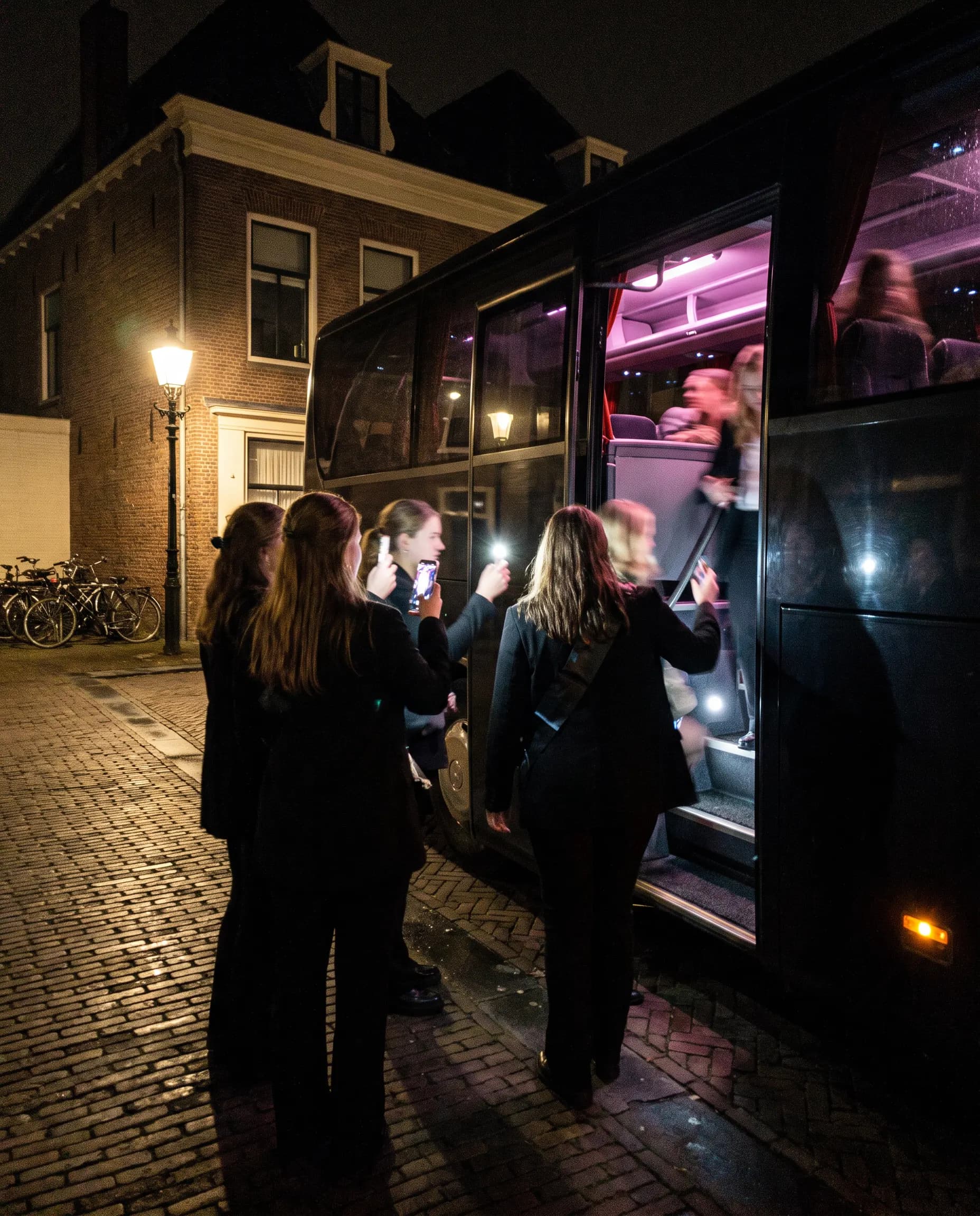 Een groep stapt 's avonds een verlichte bus in, een vrouw op de trede kijkt om naar de rest