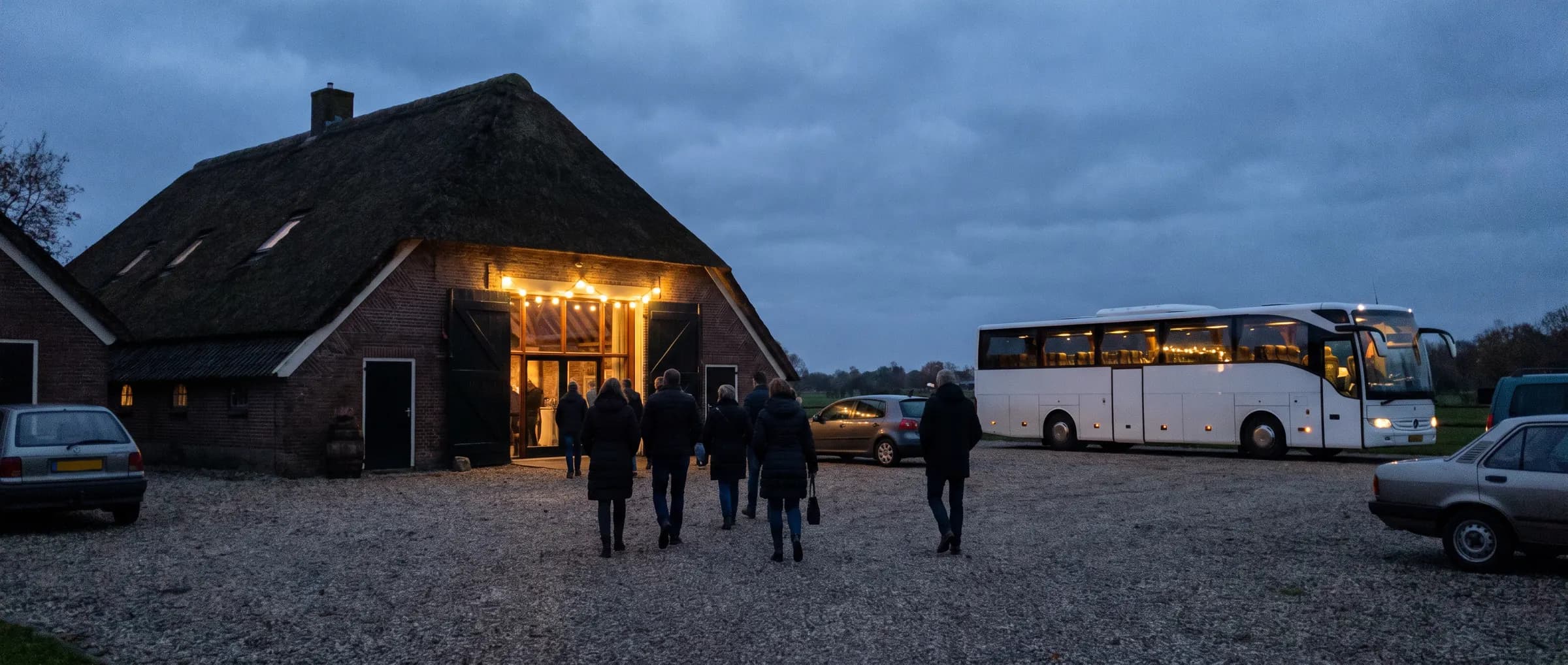 Touringcar op het erf van een boerderij met rieten dak in de schemering, gasten lopen naar de feestzaal